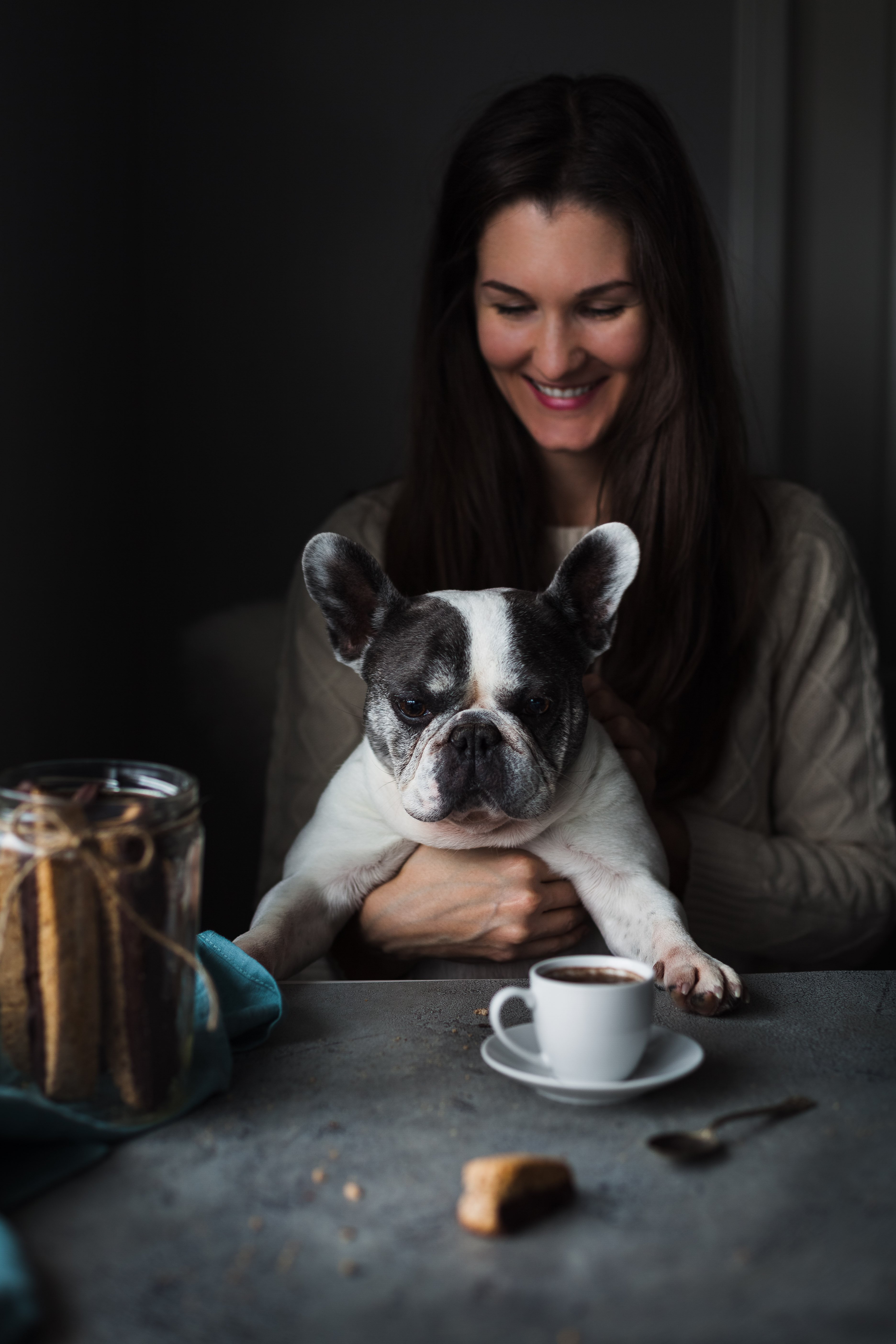 a person sitting with a white french bulldog looking at a piece of biscotti on a table.