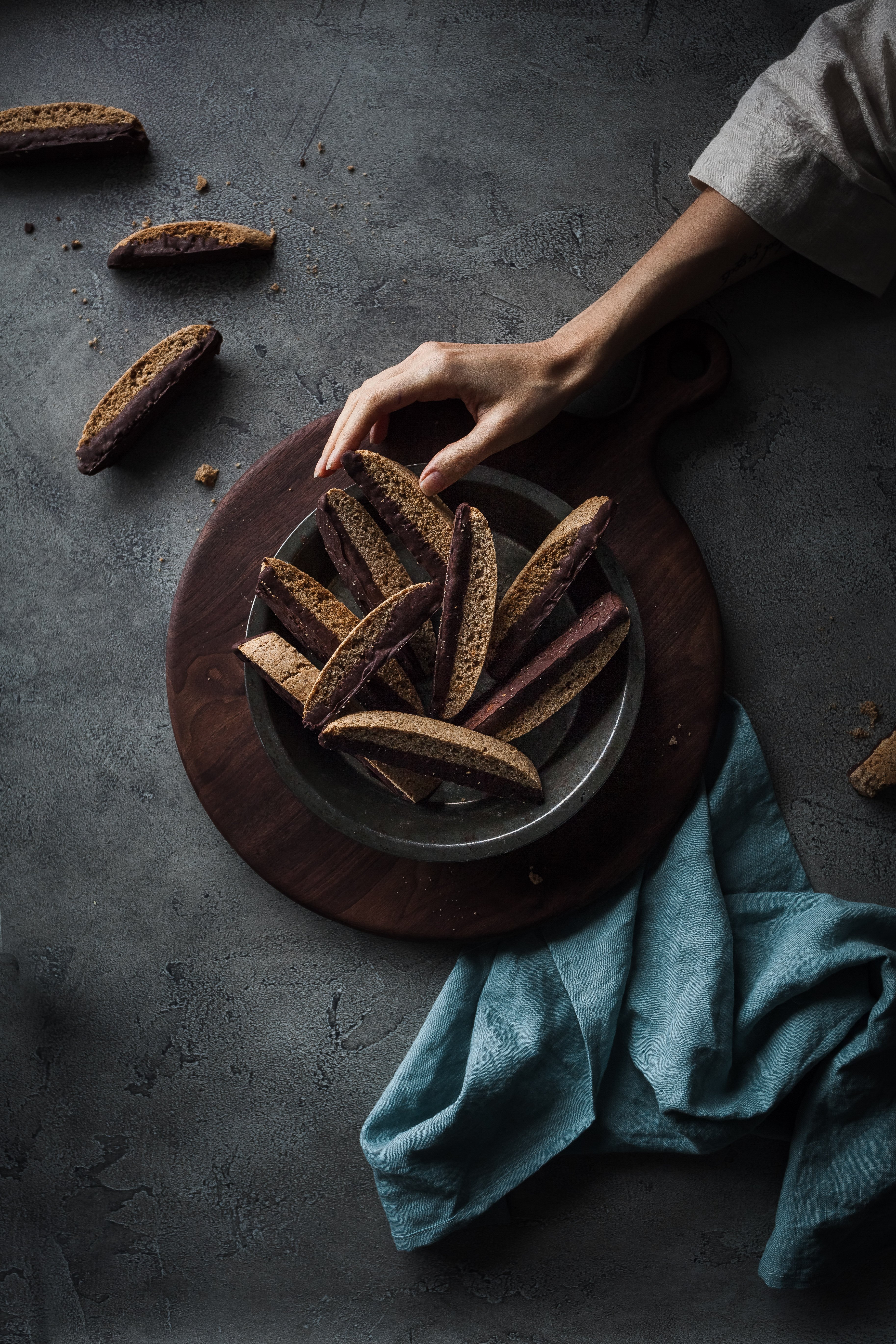 overhead image of a plate of vegan biscotti cookies on a large wooden cutting board with hand reaching for a biscotti.
