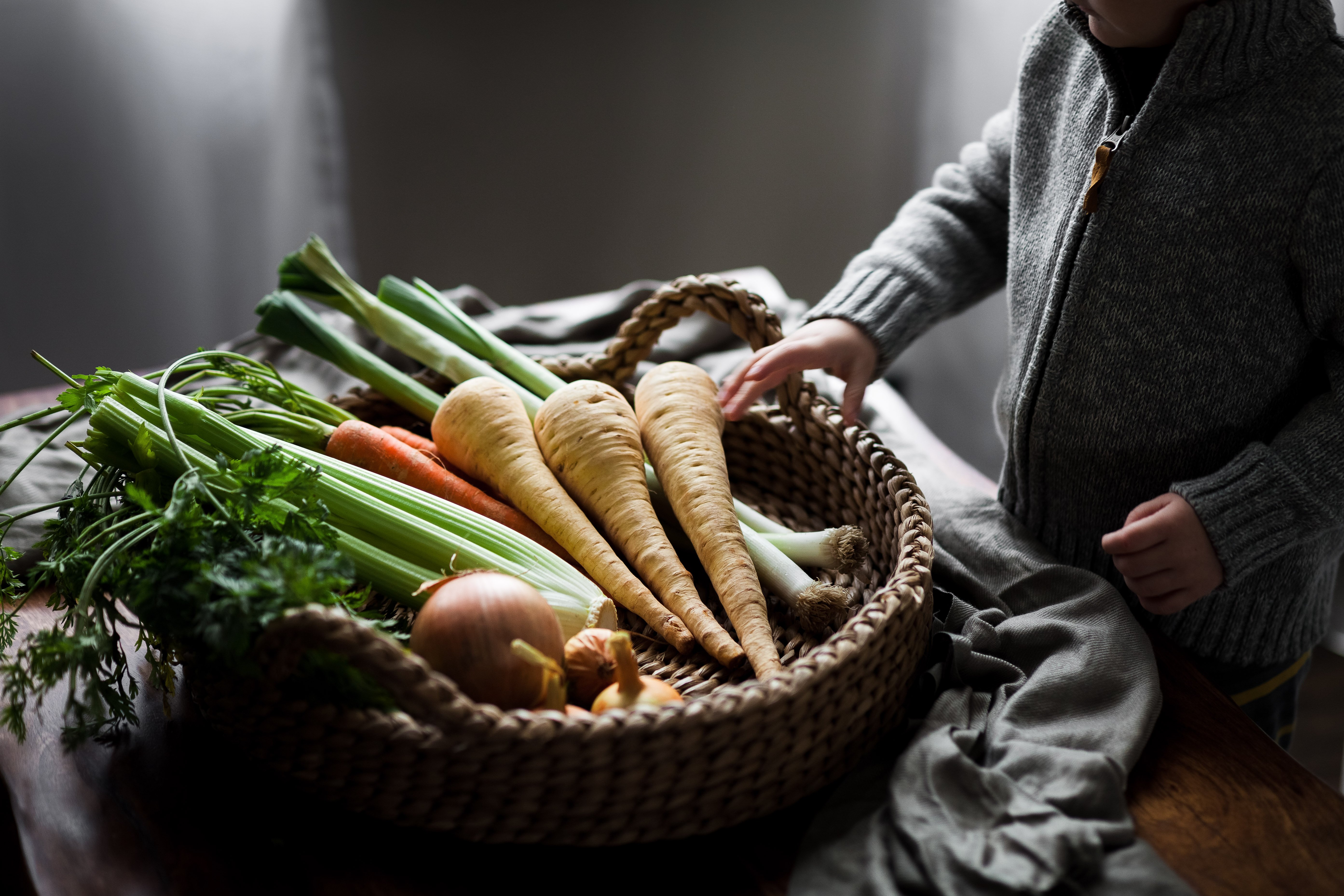 a small child grabbing root vegetables from a round basket on a wood surface.