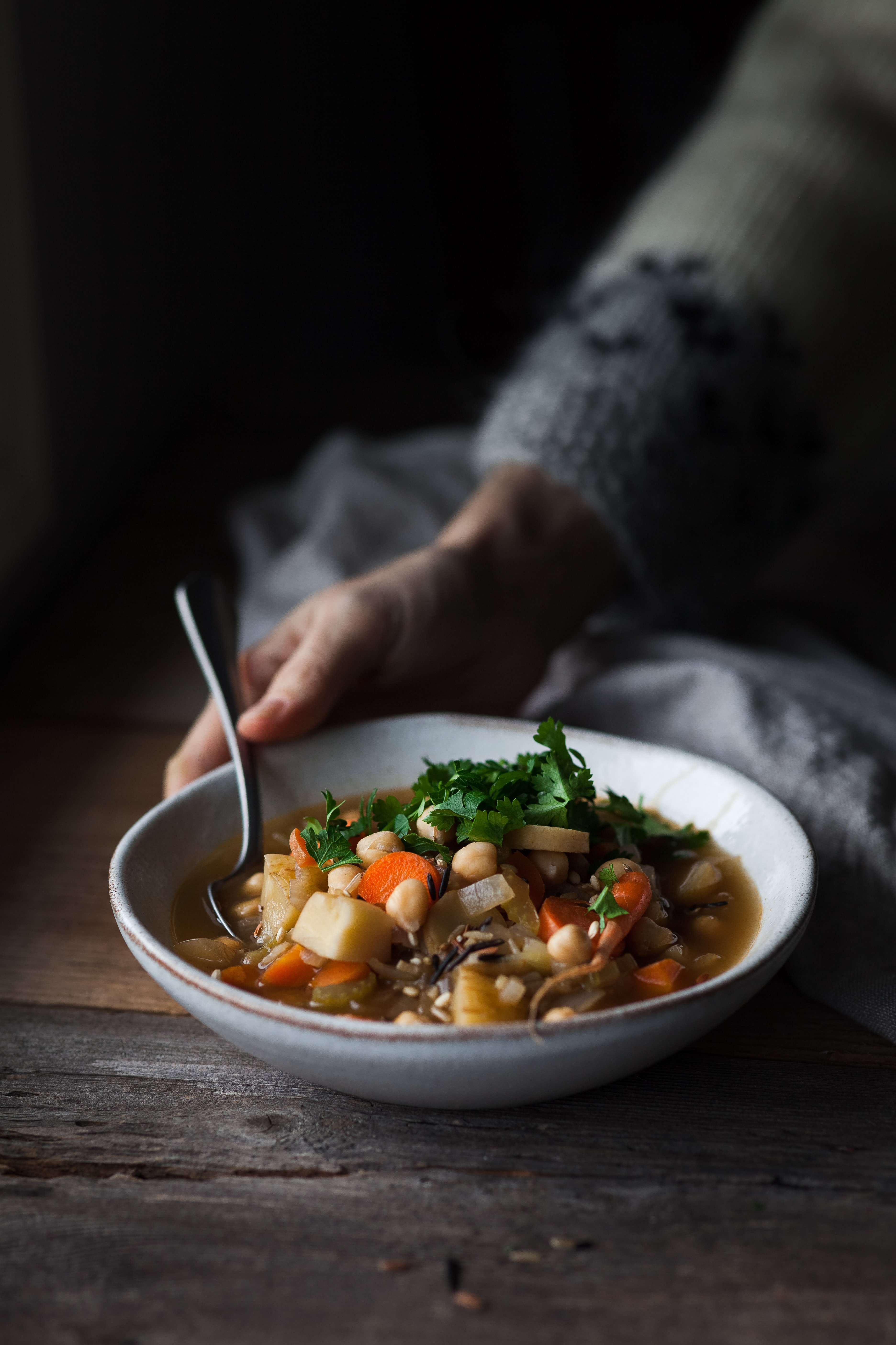 image of a bowl of winter vegetable soup with a hand holding it on a wooden surface.