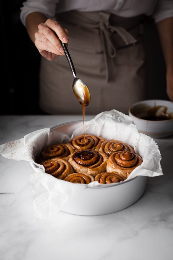 a head on image of a baker drizzling sugar glaze over Vegan Cinnamon Rolls in a pan.