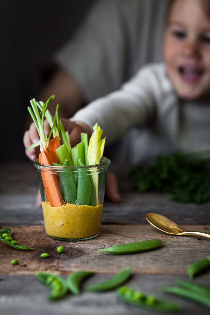 A head on image of a child reaching for cup of veggies and gold dip in a glass jar.