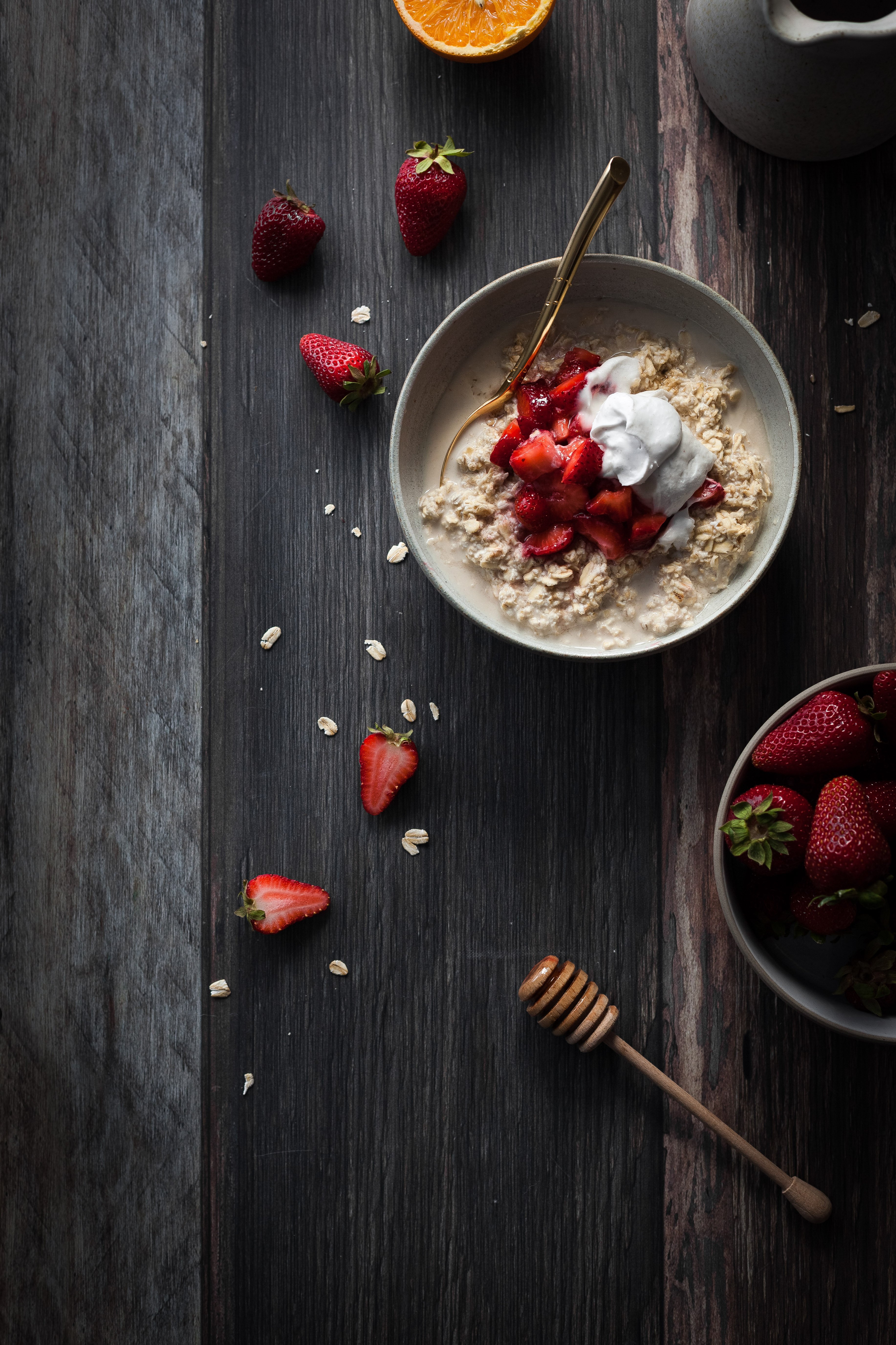overhead image of a bowl of vegan bircher museli on a wooden surface with a bowl of strawberries on the right and a honey dipper.
