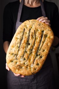 a head on image of a baker holding a loaf of focaccia bread.