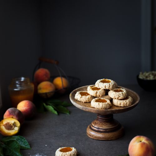 a cookie stand with thumbprint cookies on top with peaches and jam in the background.