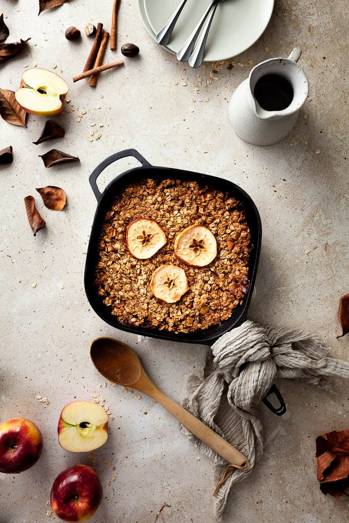 an overhead flatlay of baked oatmeal in a cast iron pan with a wooden spoon on the left and a milk jug and dishes in the upper right corner.