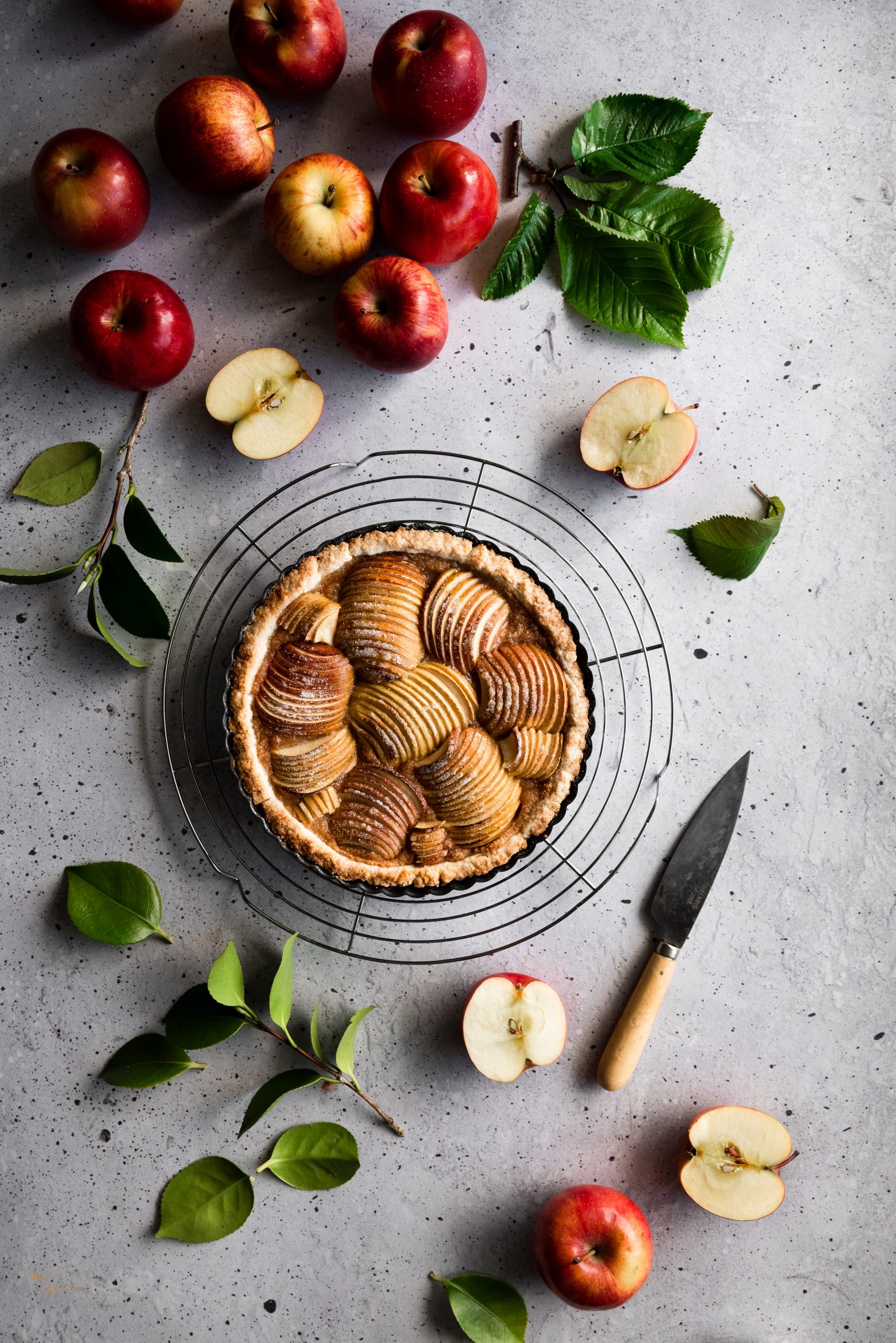 overhead image of a french apple tart on a wire rack surrounded by apples, leaves and a paring knife.