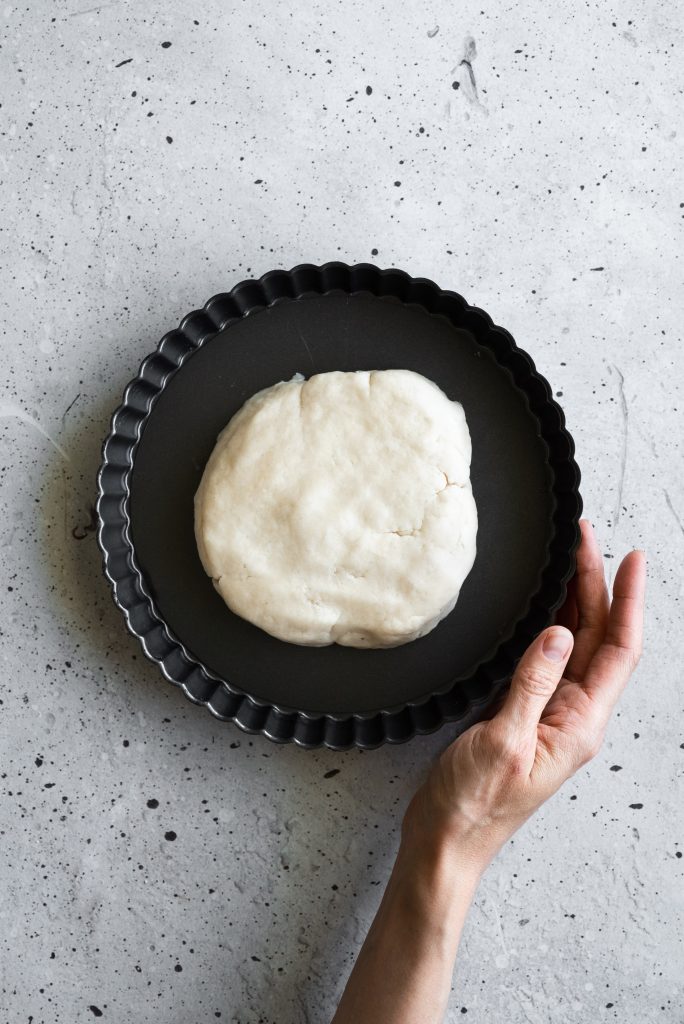 overhead image of tart dough in the middle of a tart pan with a hand holding the pan.