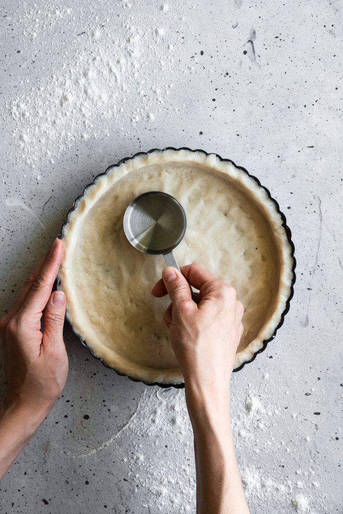 overhead image of two hands pressing tart dough into a tart pan with a small measuring cup.