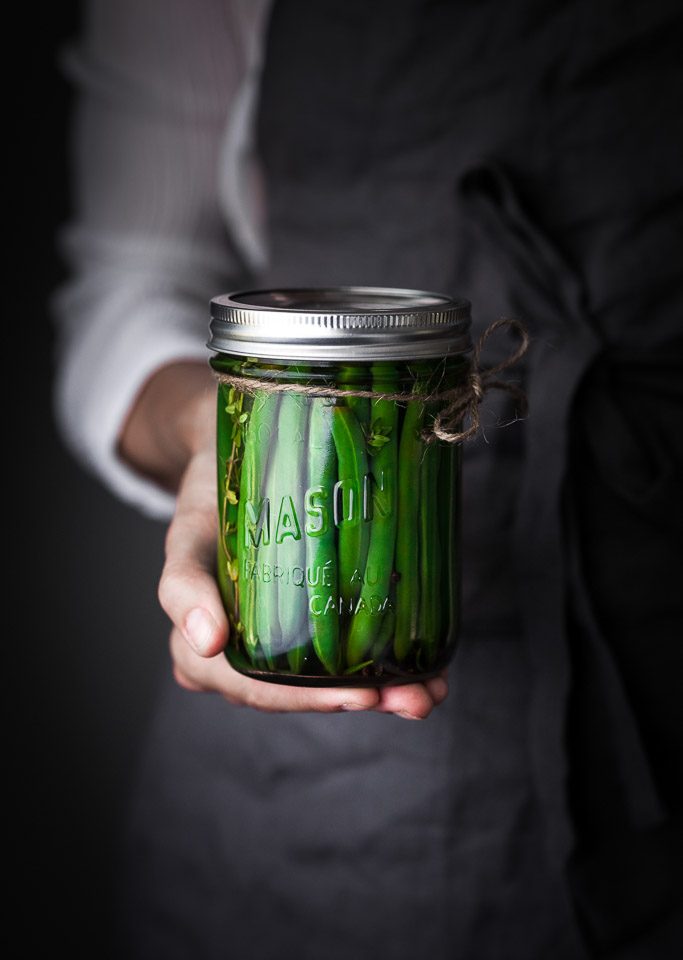 A person in a blue apron holding a jar of pickled green beans.