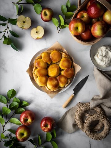 overhead view of an apple tarte tatin with a linen, apples and leaves and whipped cream on a marble surface.
