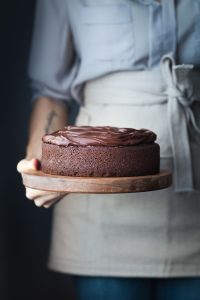 a head on image of a baker holding a chocolate cake on a wood cutting board.
