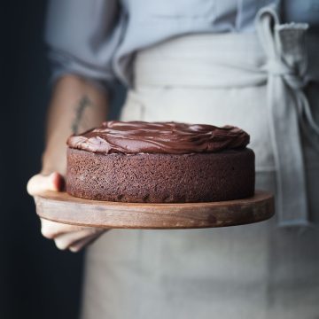a head on image of a baker holding a chocolate cake on a wood cutting board.