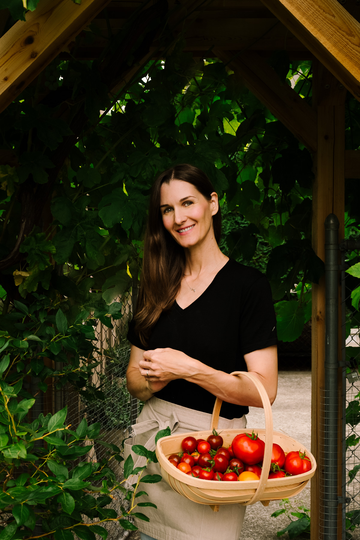A woman in jeans and an apron with a basket of tomatoes under a garden trellis.