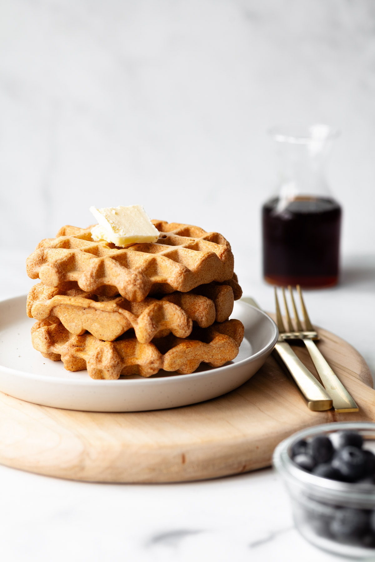 A stack of three waffles with butter on top and a carafe of maple syrup in the background with blueberries in the foreground.