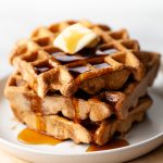 A closeup of a stack of three spelt waffles with butter and maple syrup on a white dish.