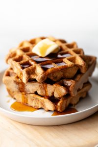 A closeup of a stack of three spelt waffles with butter and maple syrup on a white dish.
