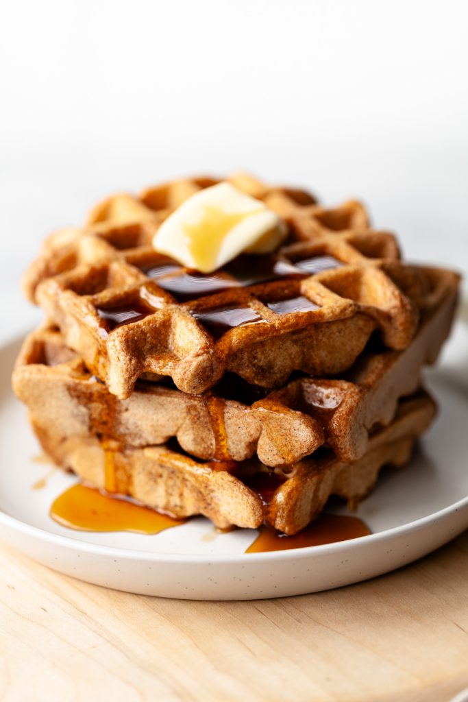 A closeup of a stack of three spelt waffles with butter and maple syrup on a white dish.