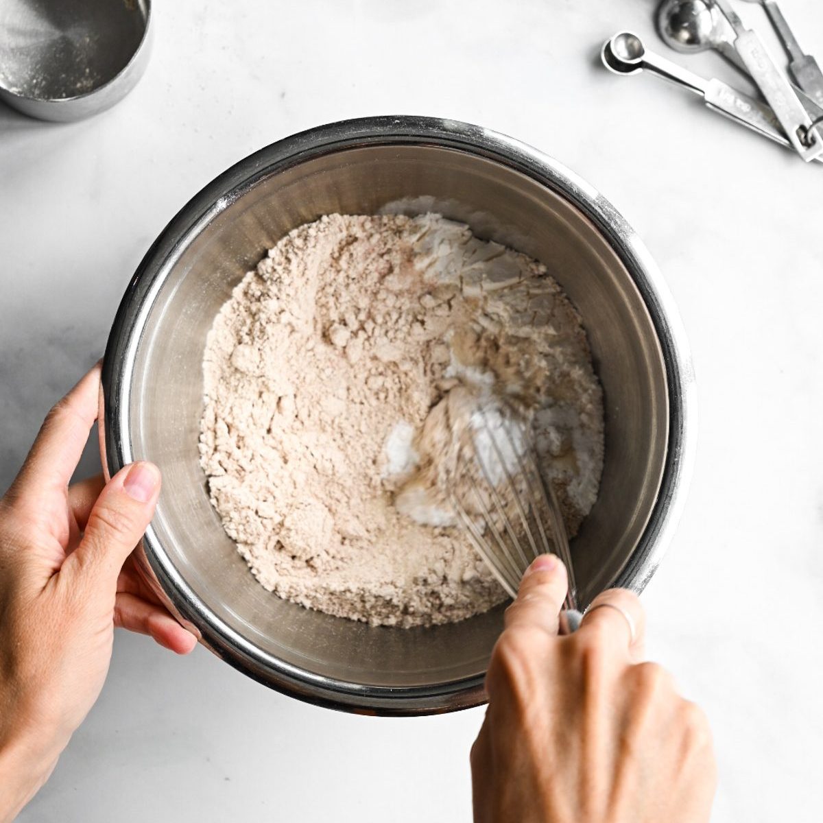 hands using a whisk to sift flour in a metal bowl.