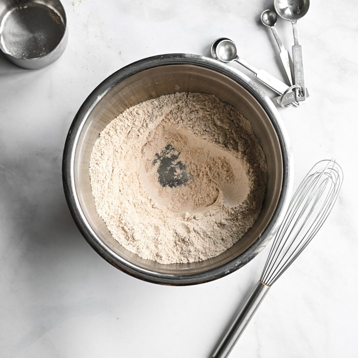 A marble surface with a bowl of flour with a well in the center and utensils on the side.