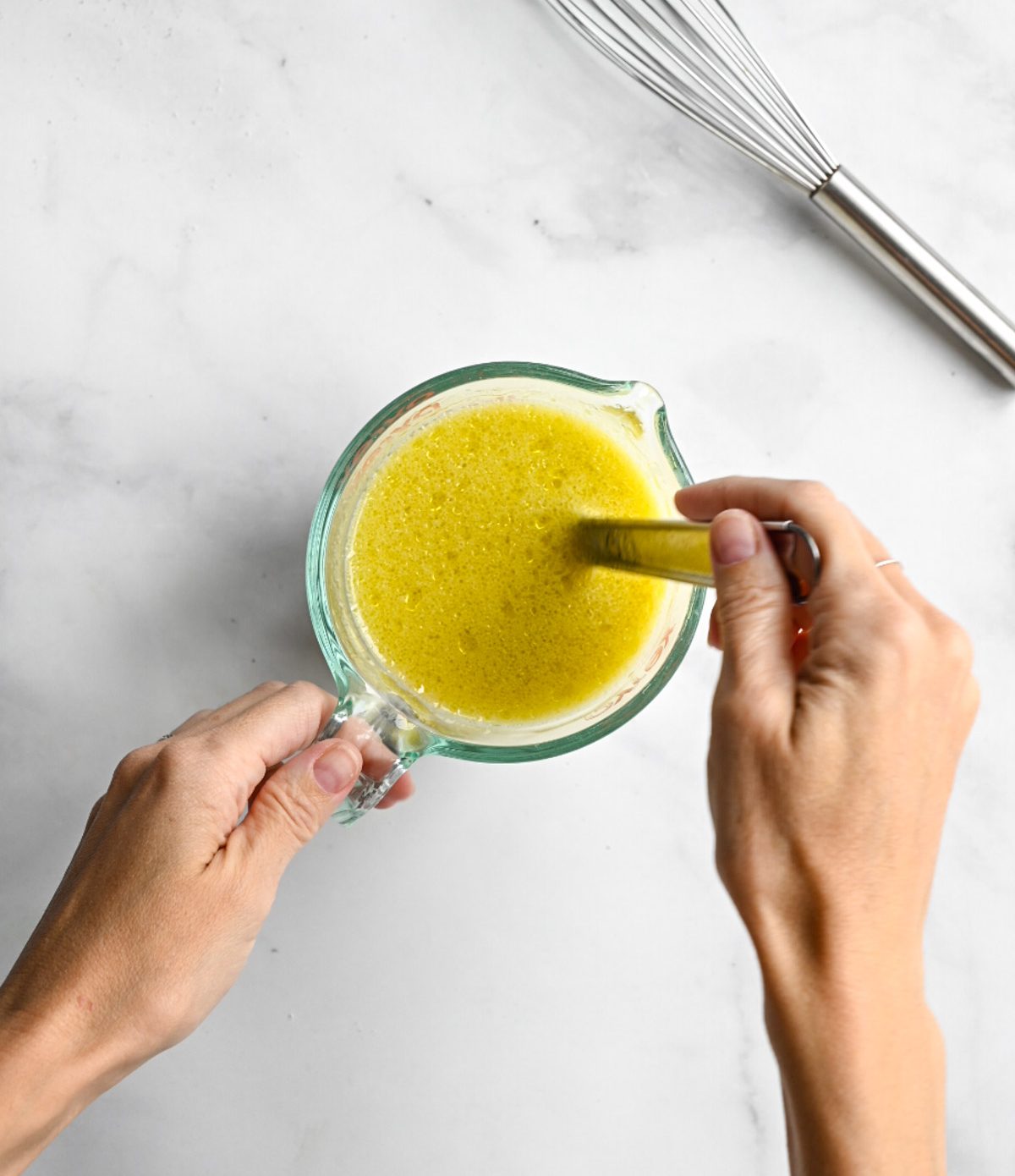 a marble surface with two hands whisking a bowl of milk and oil.
