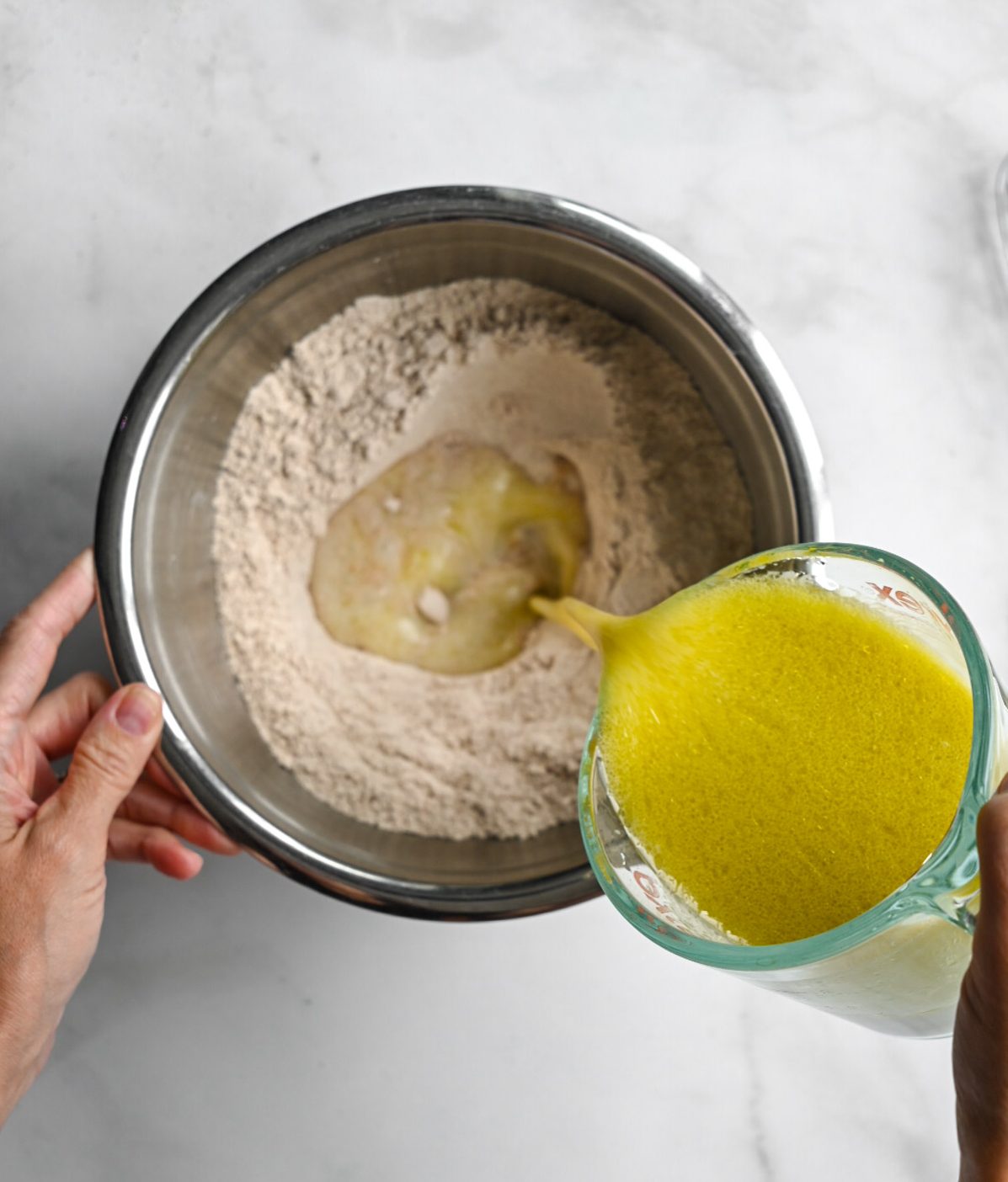 A person pouring liquids ingredients into a bowl of flour on a marble surface.