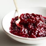 cranberry sauce in a white bowl on a white surface with a gold spoon on the side.