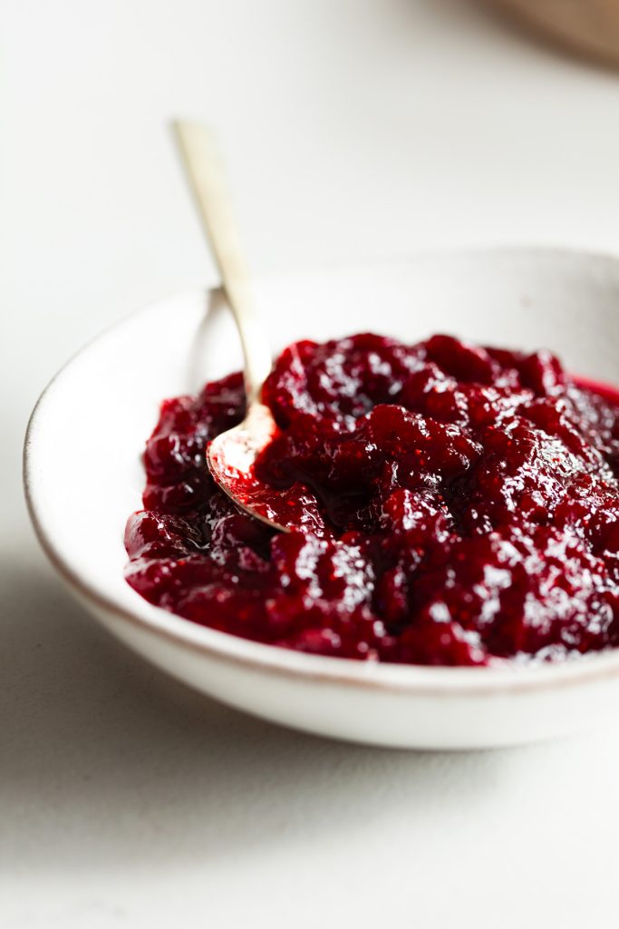 cranberry sauce in a white bowl on a white surface with a gold spoon on the side.