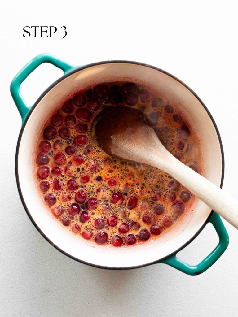 a wooden spoon stirring bubbling cranberry sauce in a blue pot on a white surface with text overlay "step 3".