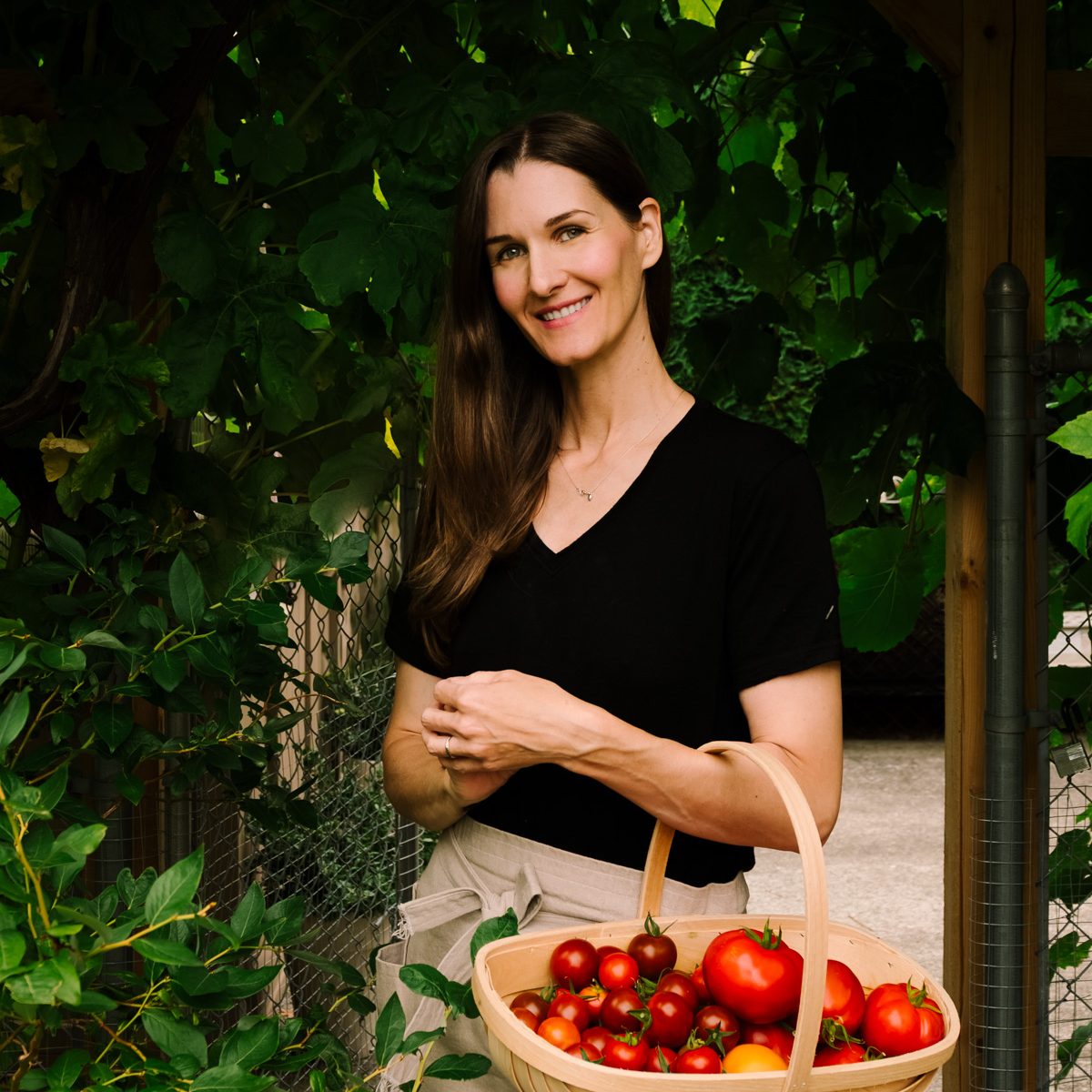 A woman in jeans and an apron with a basket of tomatoes under a garden trellis.