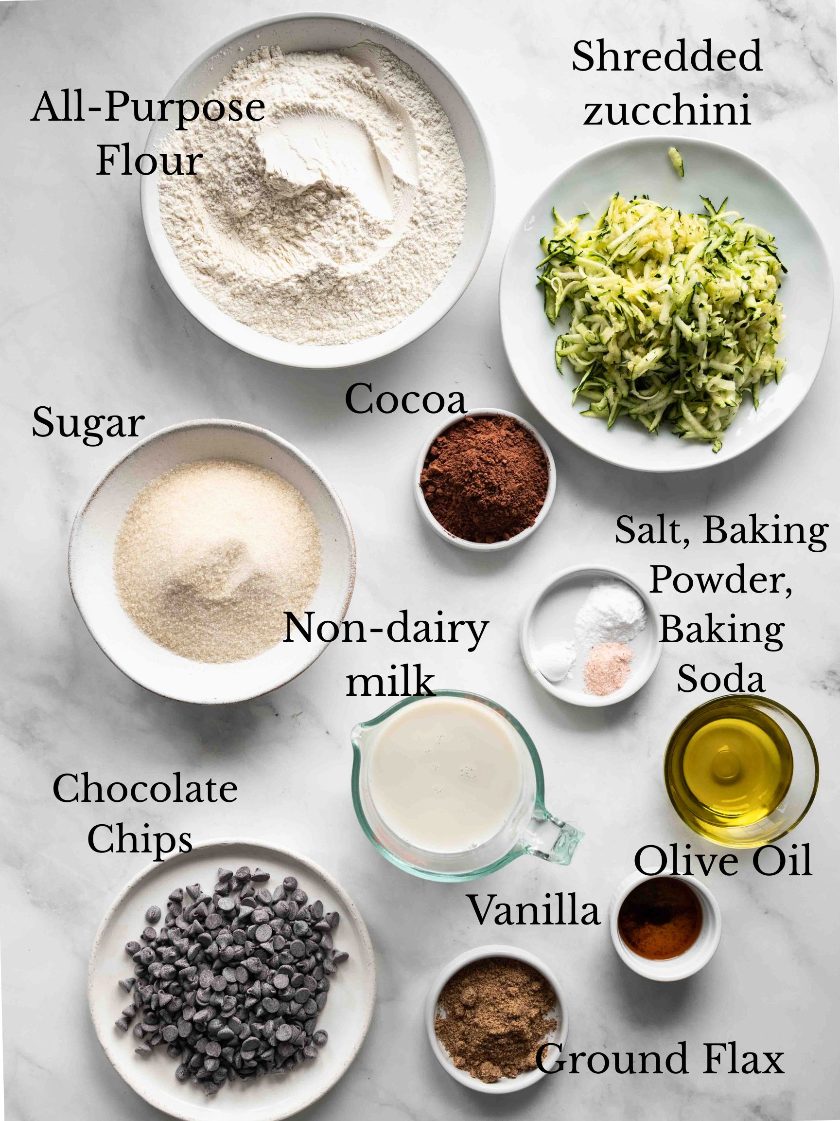 The various ingredients used to make vegan chocolate zucchini muffins on a stone surface with labels for each.