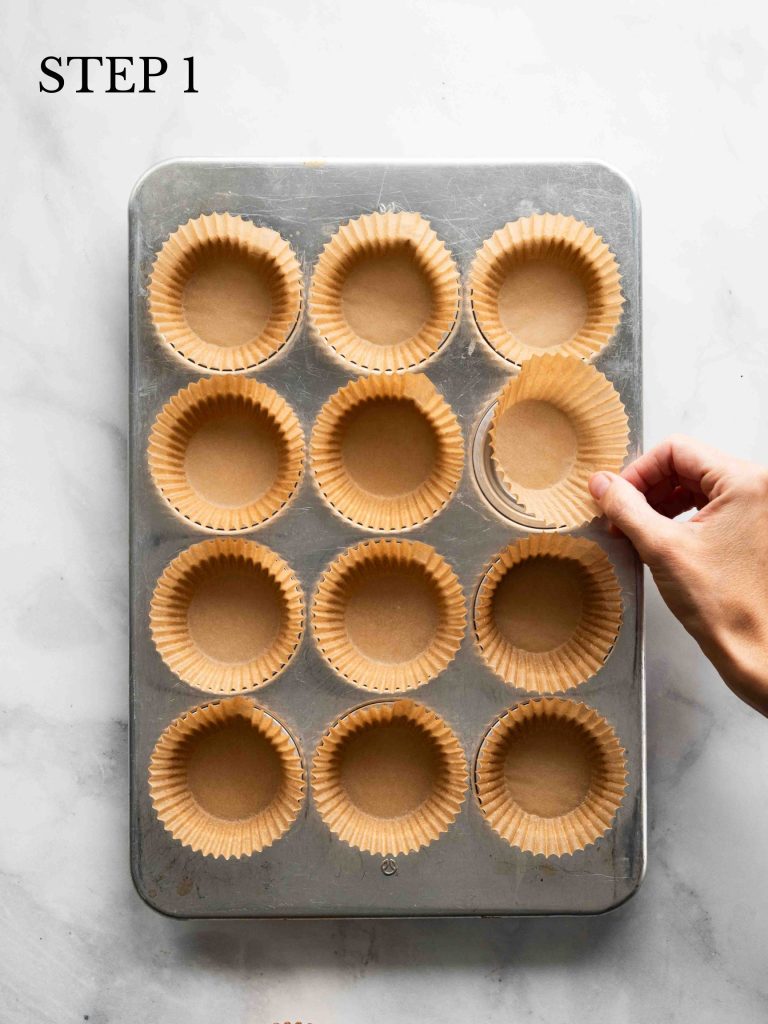 Muffin papers being placed inside a 12-cup muffin pan on a marble surface.