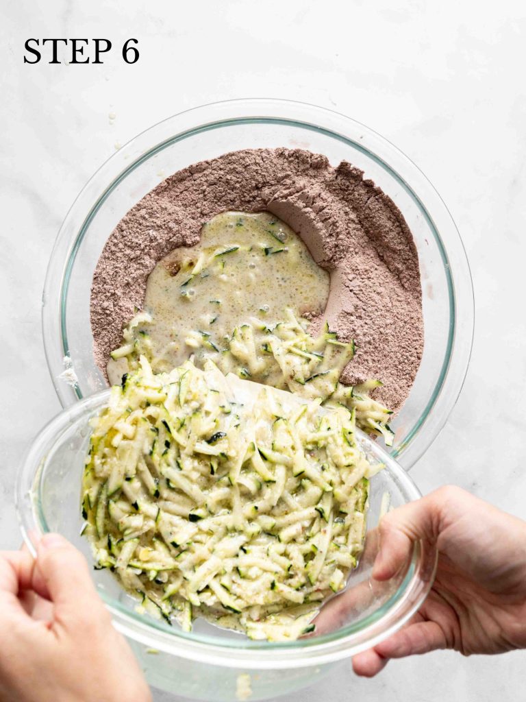 zucchini mixture being poured into dry ingredients bowl on a white surface.