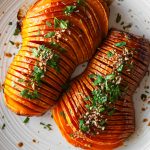 two hasselback butternut squash halves on a platter with parsley and chopped pecans.
