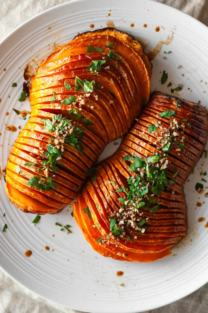 two hasselback butternut squash halves on a platter with parsley and chopped pecans.
