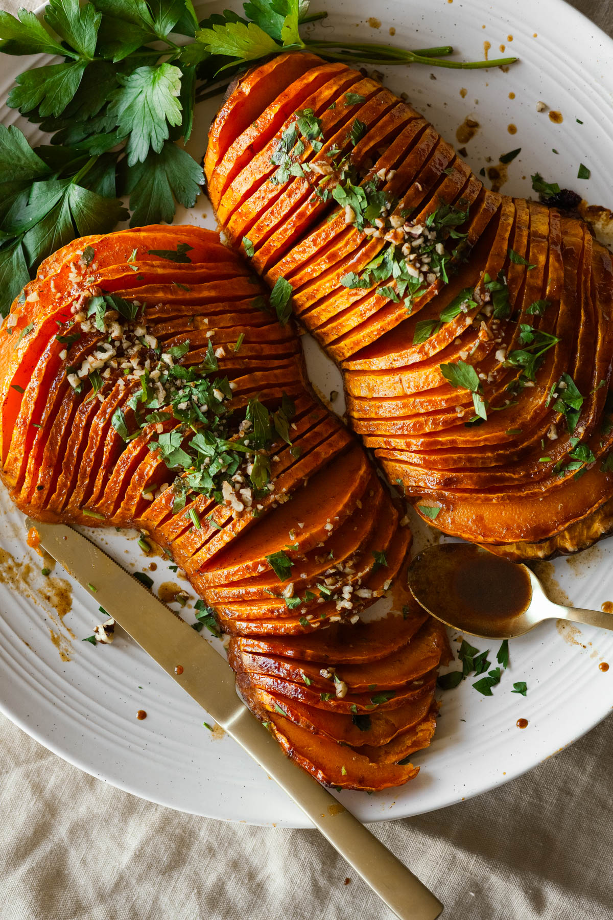 two hasselback butternut squash halves on a platter with parsley leaves, and a knife and serving spoon.