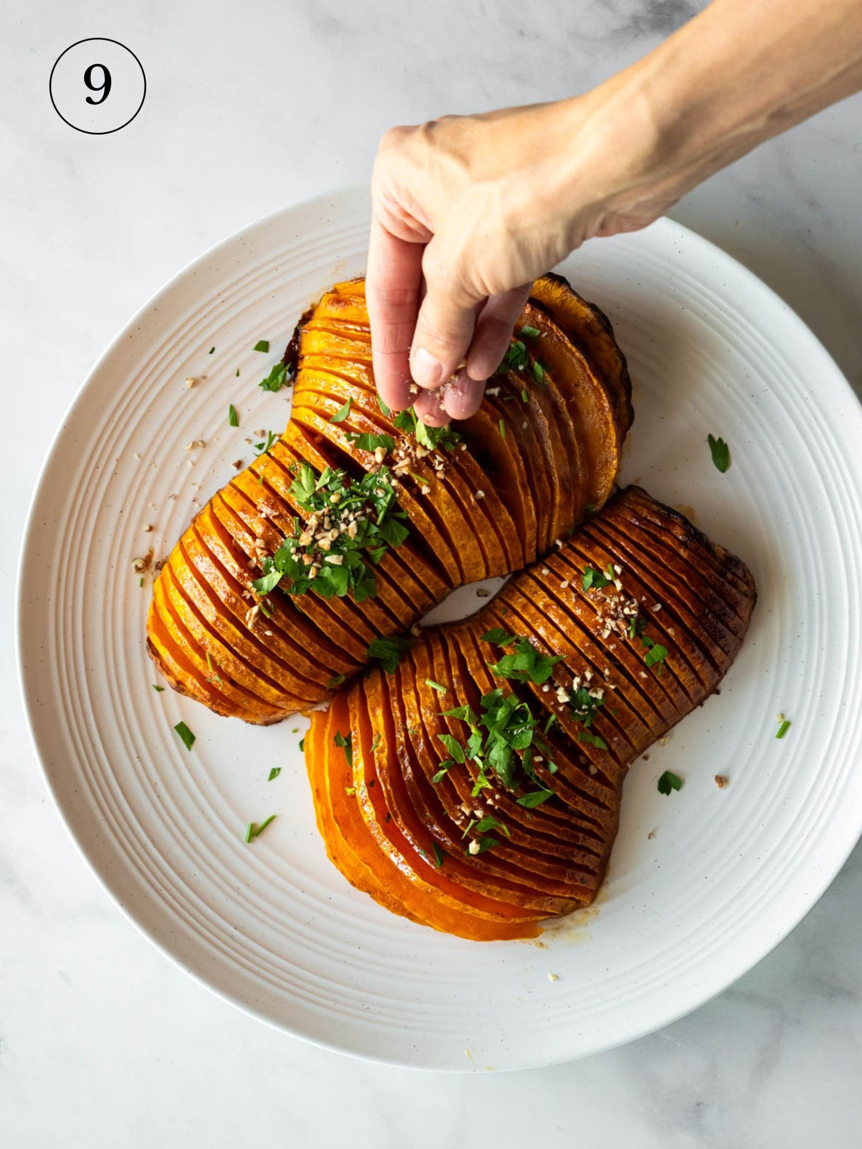a cook cprinkling parsley on top baked hasselback squash on a white serving dish.