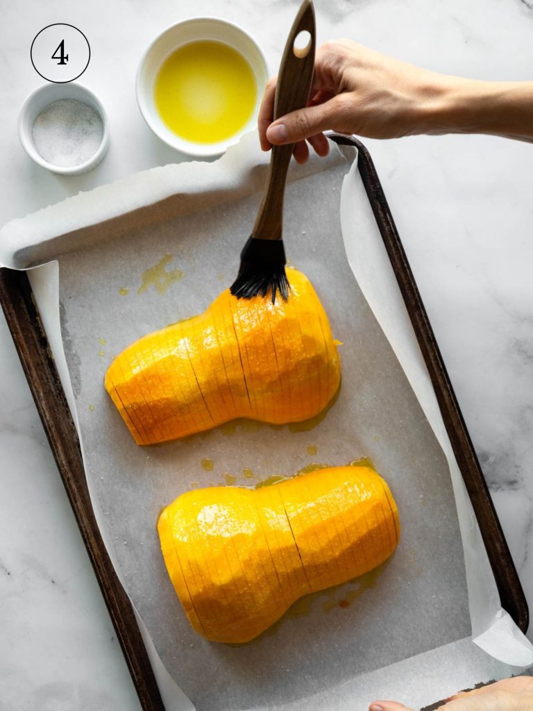 basting olive oil on the top of two hasselback squash halves on a baking sheet.